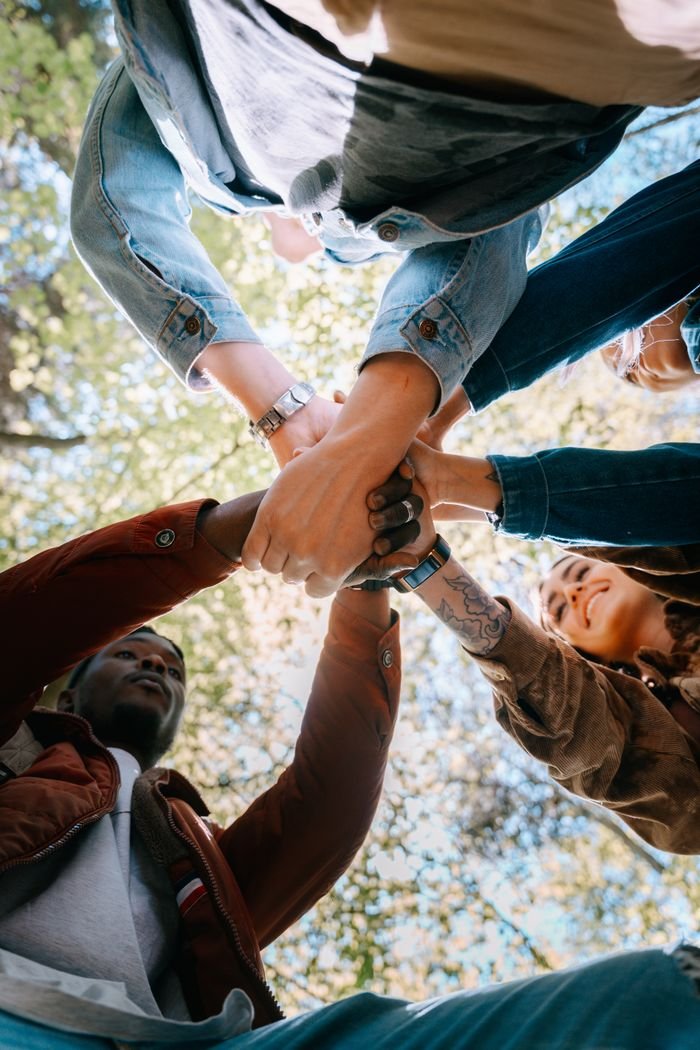 A diverse group of adults joining hands outdoors, symbolizing unity and teamwork from a low angle perspective.